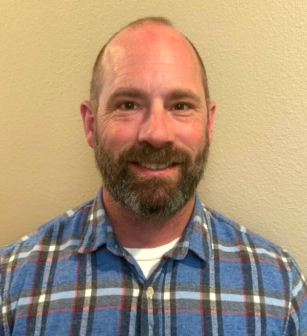 Man with a short beard and balding head smiles at the camera, wearing a blue plaid shirt against a beige wall.