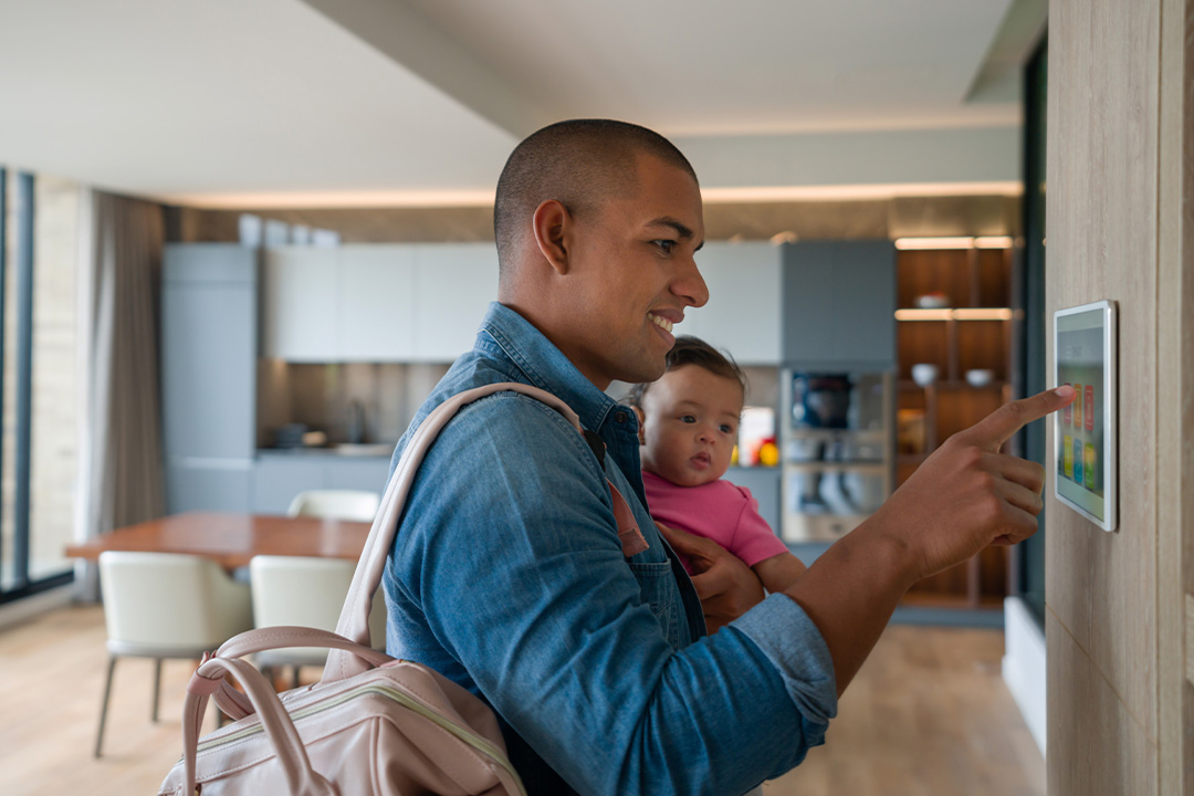 Man holding a baby while tapping a wall-mounted touchscreen control panel in a modern kitchen/dining area.