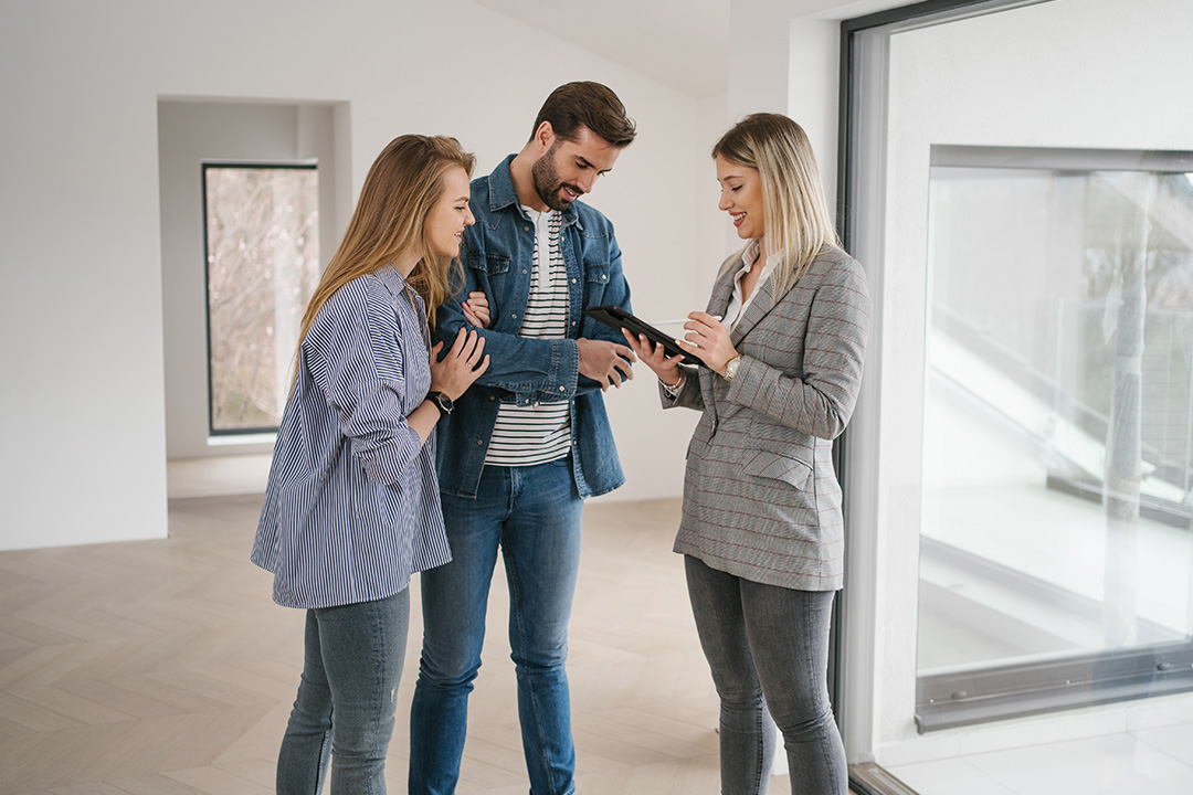 Three adults stand in a bright, empty living space looking at a tablet together and smiling, as if reviewing a property. -OR- They are discussing a home plan in a vacant modern room.