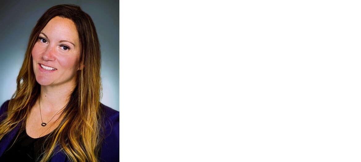 Portrait of a smiling woman with long brown hair, wearing a dark blazer against a gray backdrop, suitable as a professional headshot.